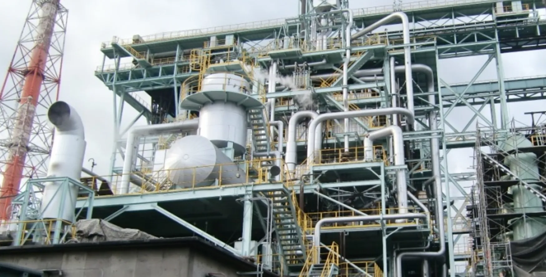 緻密に組み上げられたプラントの銀色の配管群と蒸気、背後にそびえる紅白の煙突。丸栄産業が手掛ける防食メンテナンスの主要な現場風景