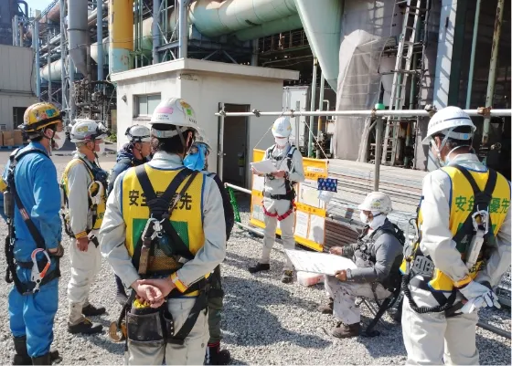 工場の屋外で、安全ベストとヘルメットを着用した作業員たちが集まって打ち合わせをしている様子。