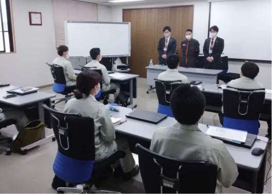 会議室で作業服を着た社員たちが、前方に立つ講師や担当者の話を座って聞いている様子。