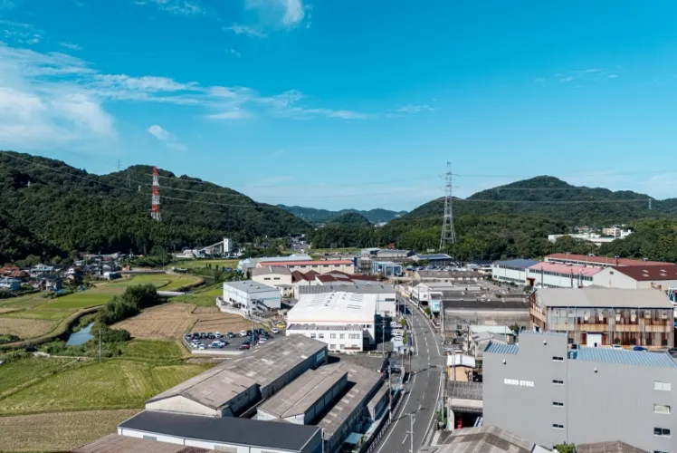 青空の下、緑豊かな山と田畑に隣接する、工場や倉庫が立ち並ぶエリアを上空から捉えた写真
