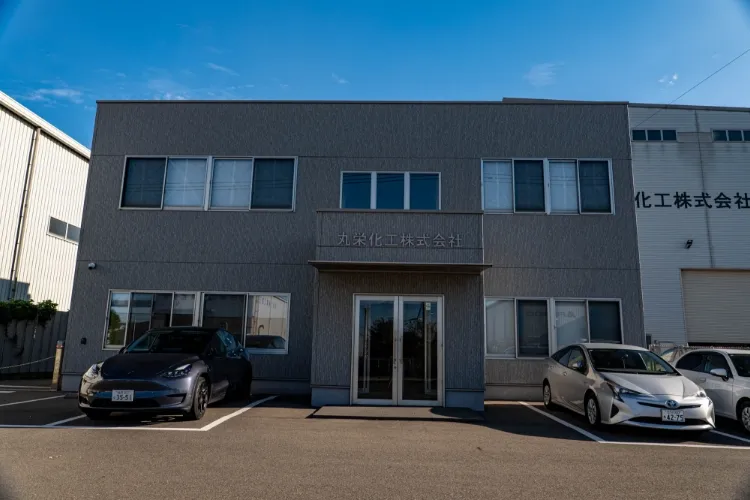 正面に「丸栄化工株式会社」と掲げられた、グレーの2階建て社屋とその前の駐車場。